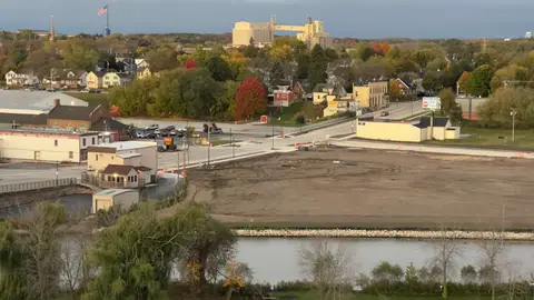 Fall color, a flag up high, a tall pale industrial building, and a cleared lot by the water.