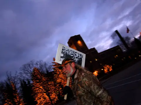 A lone picketer with a sign at dusk, factory glow behind, outside Kohler Co. headquarters.