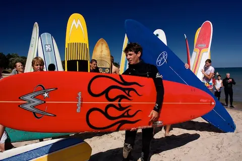 Surfer hauling a flame-painted board past a rack of longboards.
