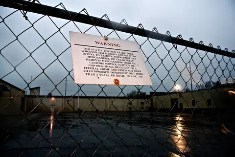 Chain-link fence with a warning sign over a wet, empty industrial lot at the Sheboygan Paper Box site.