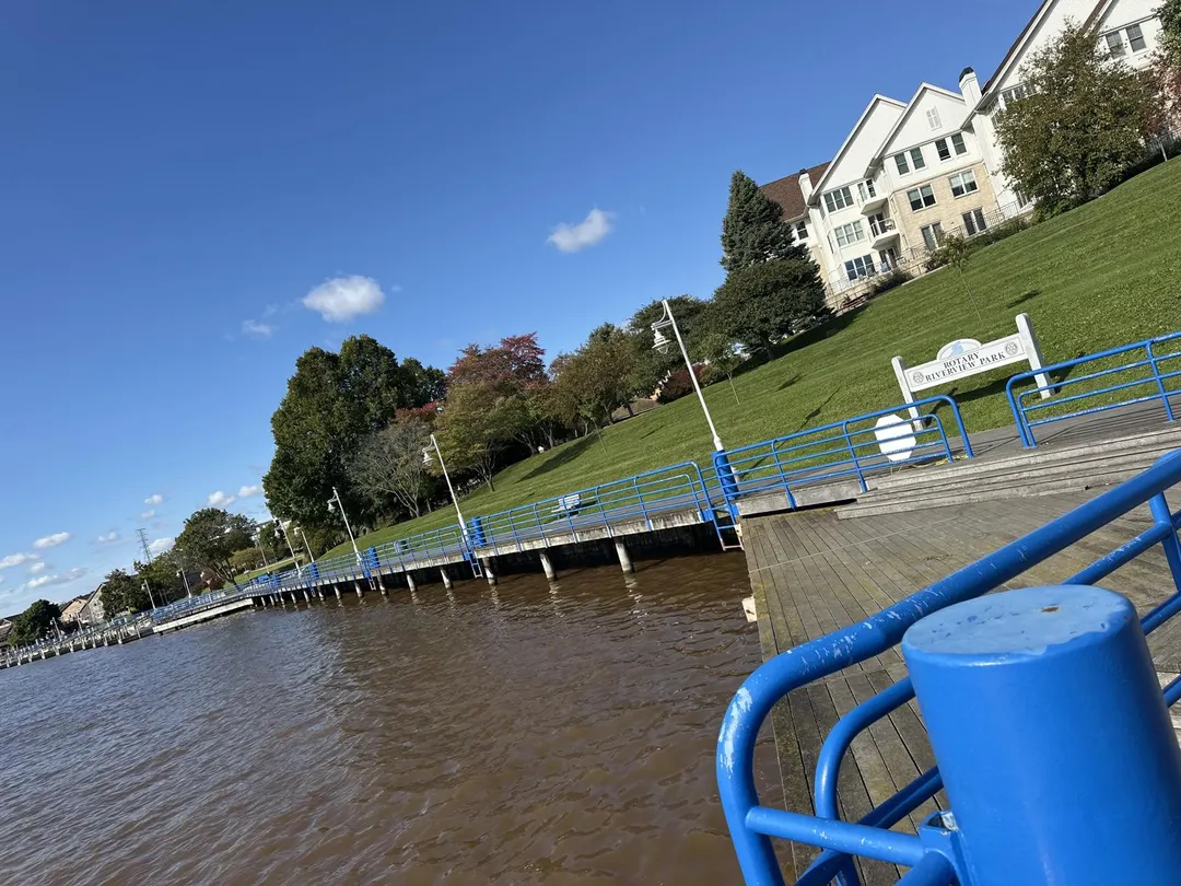 The riverwalk at Rotary Riverview Park: blue railings, a wooden boardwalk along brown river water, the park sign on a grassy hill and white buildings above. No people in the photo.