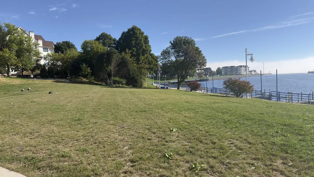 An open green lawn beside the Sheboygan River, with a blue railing along the water and a white lamppost. No people in the photo.